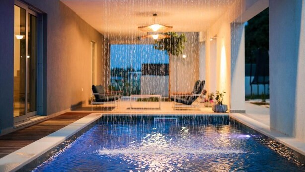 Una piscina moderna al coperto con cascata, circondata da poltrone relax e un ventilatore a soffitto.