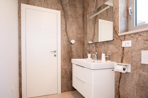 Ein Badezimmer mit weißer Tür, einer Marmorwand, einem Waschbecken mit Spiegel darüber und einem Toilettenpapierhalter.