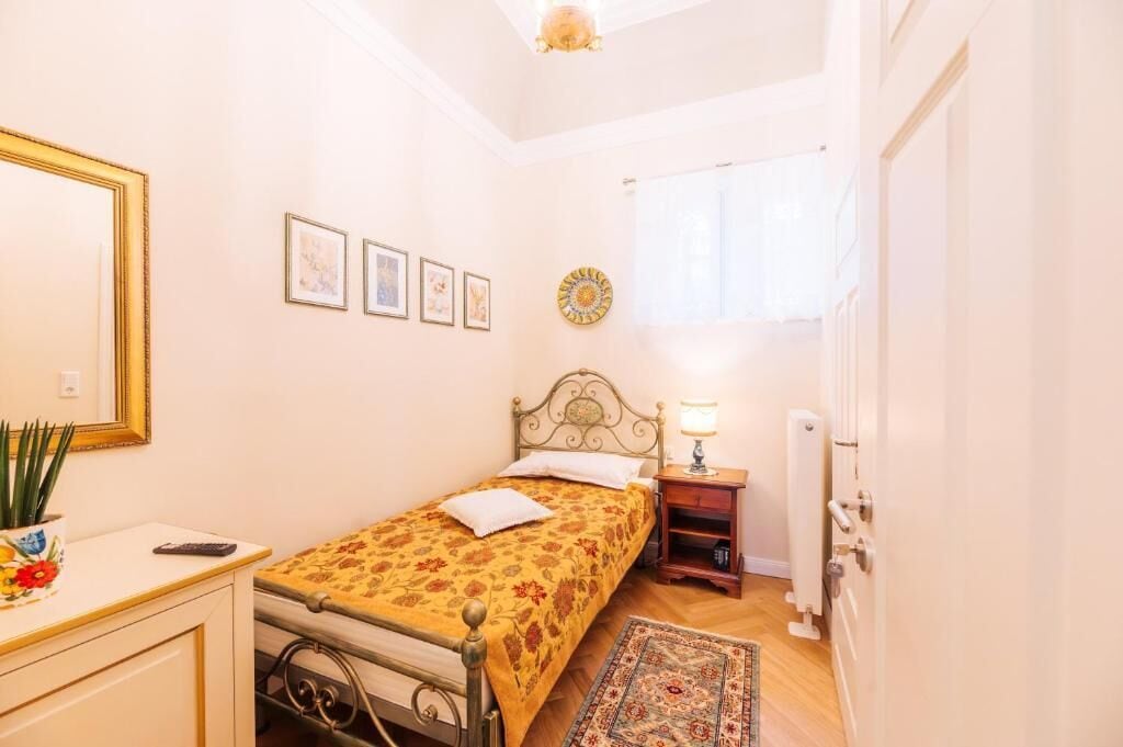 A small, well-lit bedroom with a bed, bedside table, lamp, and a decorative wall piece.