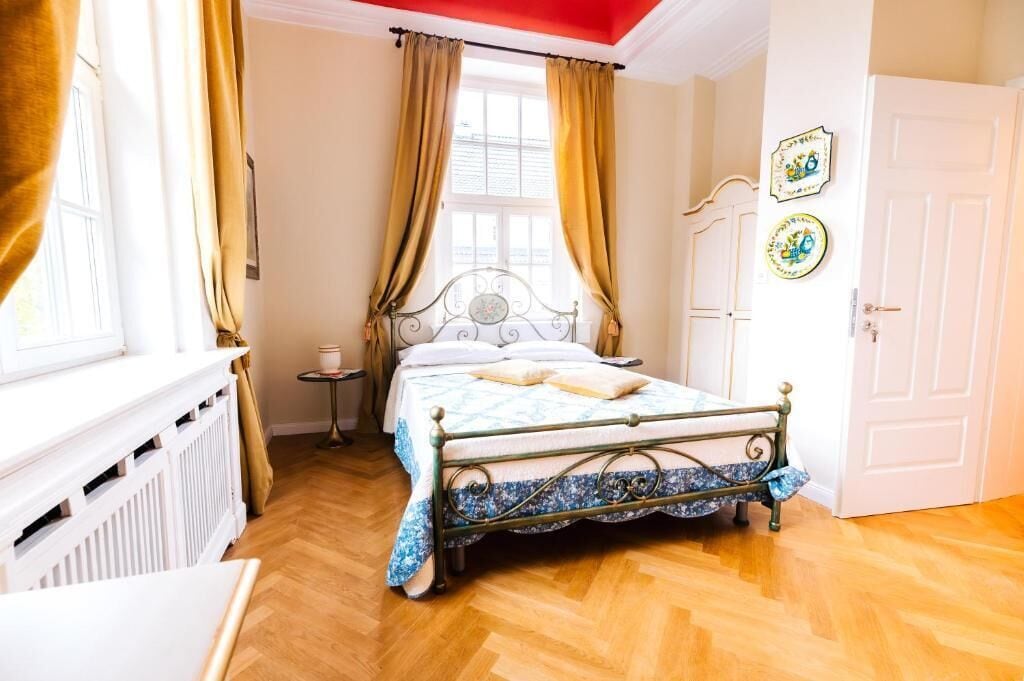 Un dormitorio con estructura de cama metálica, radiador blanco, ventana con cortinas y una puerta.