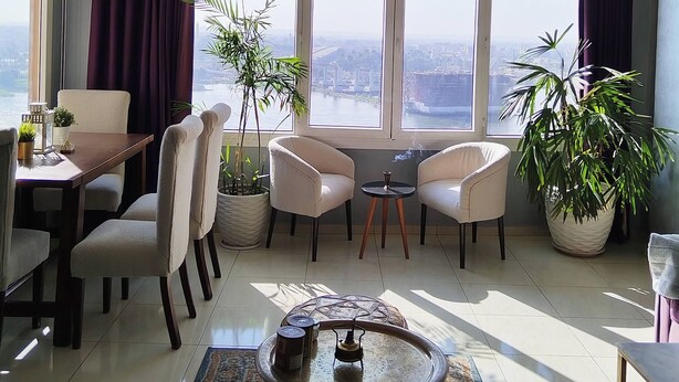 A spacious living room with a high ceiling, large windows, and a view of the city. There are two armchairs, a coffee table, and a dining area with a table and chairs. - The Traveler Nile and Pyramid View Magical Sunrises, Historic Wonders (Cairo)