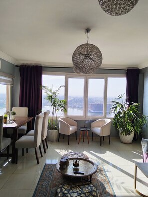 A spacious living room with a high ceiling, large windows, and a view of the city. There are two armchairs, a coffee table, and a dining area with a table and chairs. - The Traveler Nile and Pyramid View Magical Sunrises, Historic Wonders (Cairo)