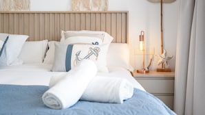 A neatly made bed with a blue bedspread, white pillows, and a rolled white towel. There is a wooden headboard, a wall-mounted light fixture, and a small wooden nightstand with a lamp and a starfish decoration. - Modern 3-pax Apartment With Solarium (Casares)