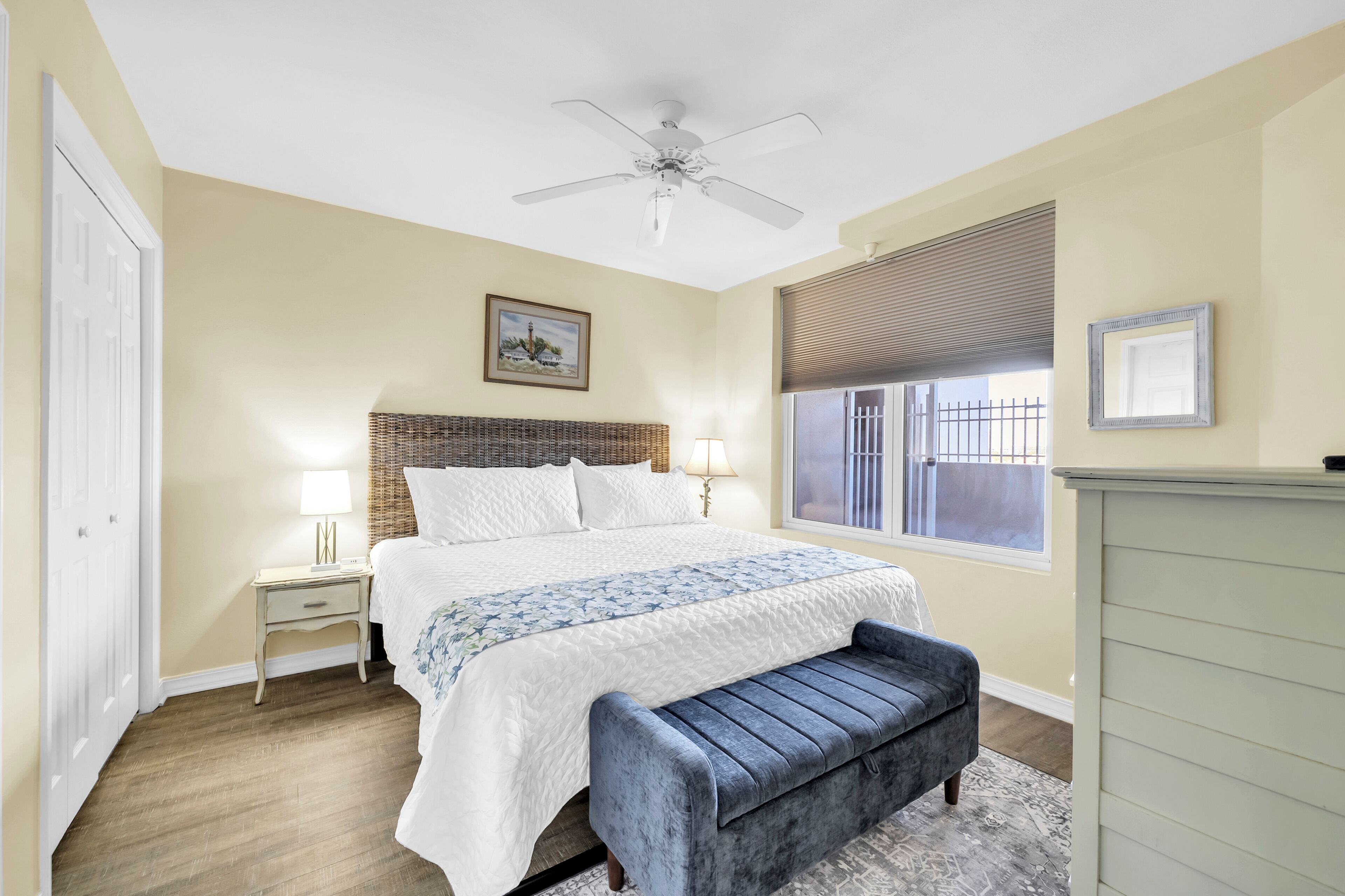 A bedroom with a bed, bedside tables, a bench, a ceiling fan, a window with blinds, and a picture on the wall.