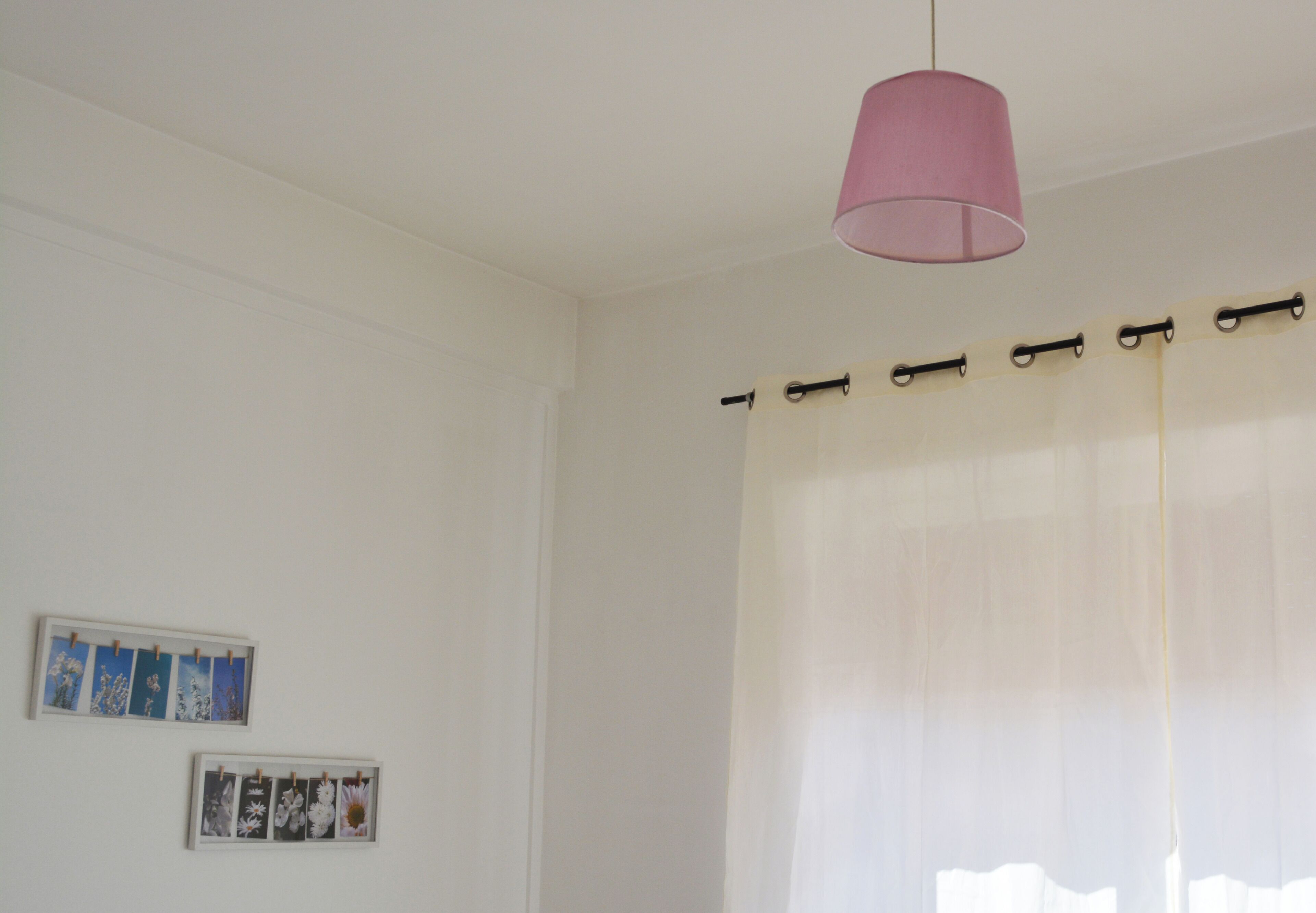 Een kamer met een witte muur, een roze hanglamp en een raam met dunne gordijnen.