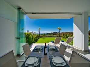 An outdoor dining area with a table set for two, overlooking a landscaped garden and a clear blue sky. - Crystal Lagoon Apartment Retreat (Casares)
