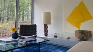 Un salon moderne avec une table basse en verre, un fauteuil en cuir, une lampe et une vue sur la forêt grâce à de grandes fenêtres.