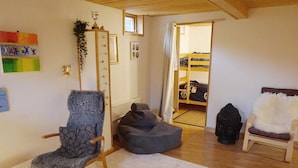 Um quarto acolhedor com soalho de madeira, uma poltrona cinzenta, uma cadeira branca e um pequeno sofá. Há um armário de madeira com uma planta em cima e uma janela com cortinas.