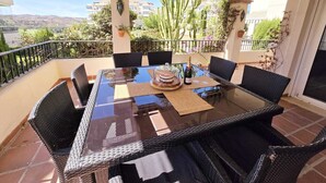 Un tavolo da pranzo con piano in vetro, sedie in vimini, una torta, una bottiglia di champagne e sullo sfondo edifici e cactus.