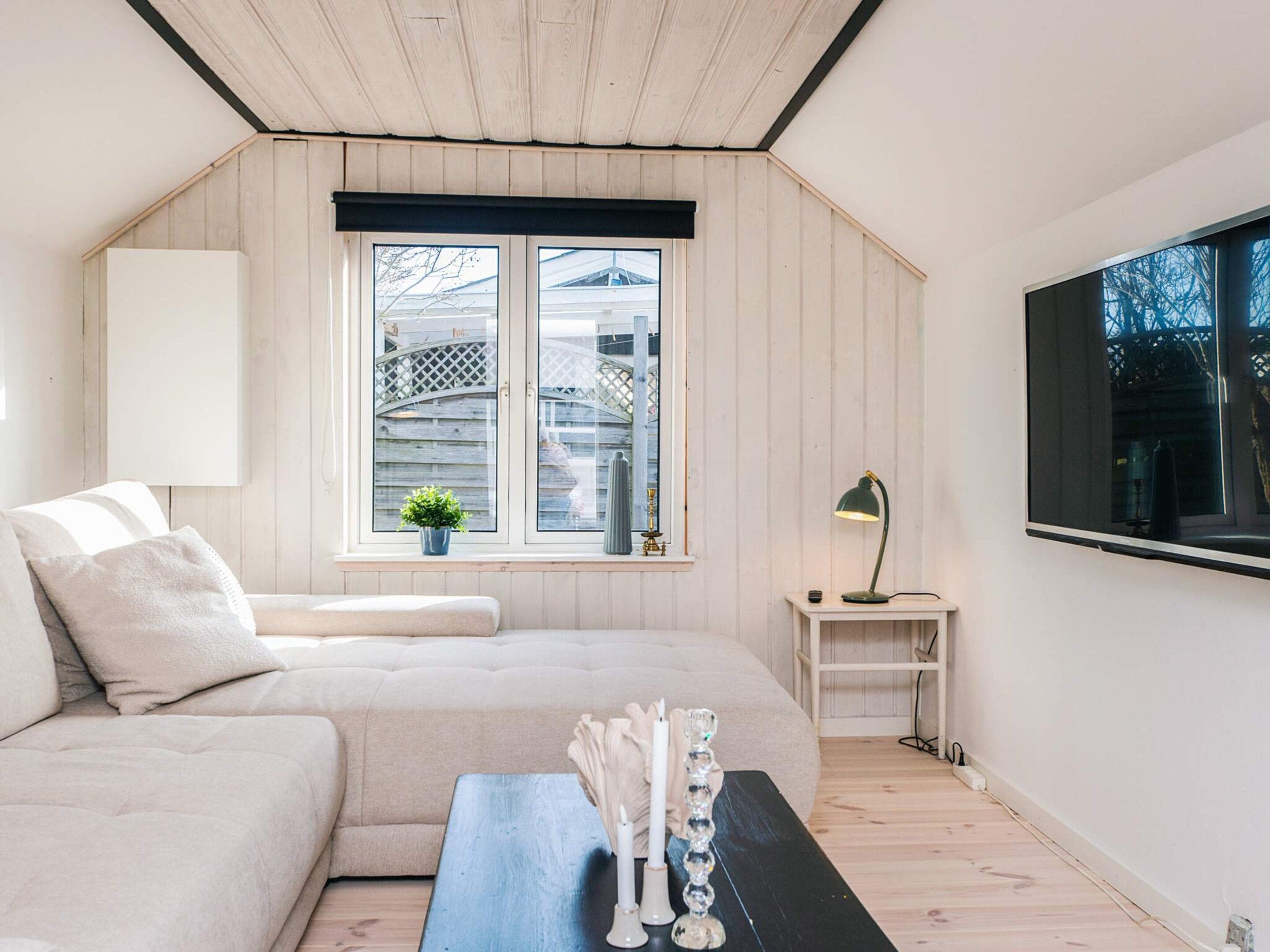 Un dormitorio moderno y minimalista con techo inclinado, un ventanal grande, un sofá, una mesita de noche con velas y un televisor montado en la pared.