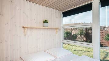 A bedroom with a wooden ceiling, a bed with white bedding, a small shelf with a plant, and a window with a view of the outdoors.