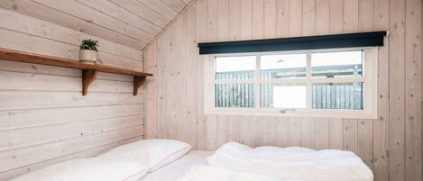 A bedroom with a wooden slatted ceiling, a window with a view of a fence, a bedside shelf with a plant, and a bed with white bedding.