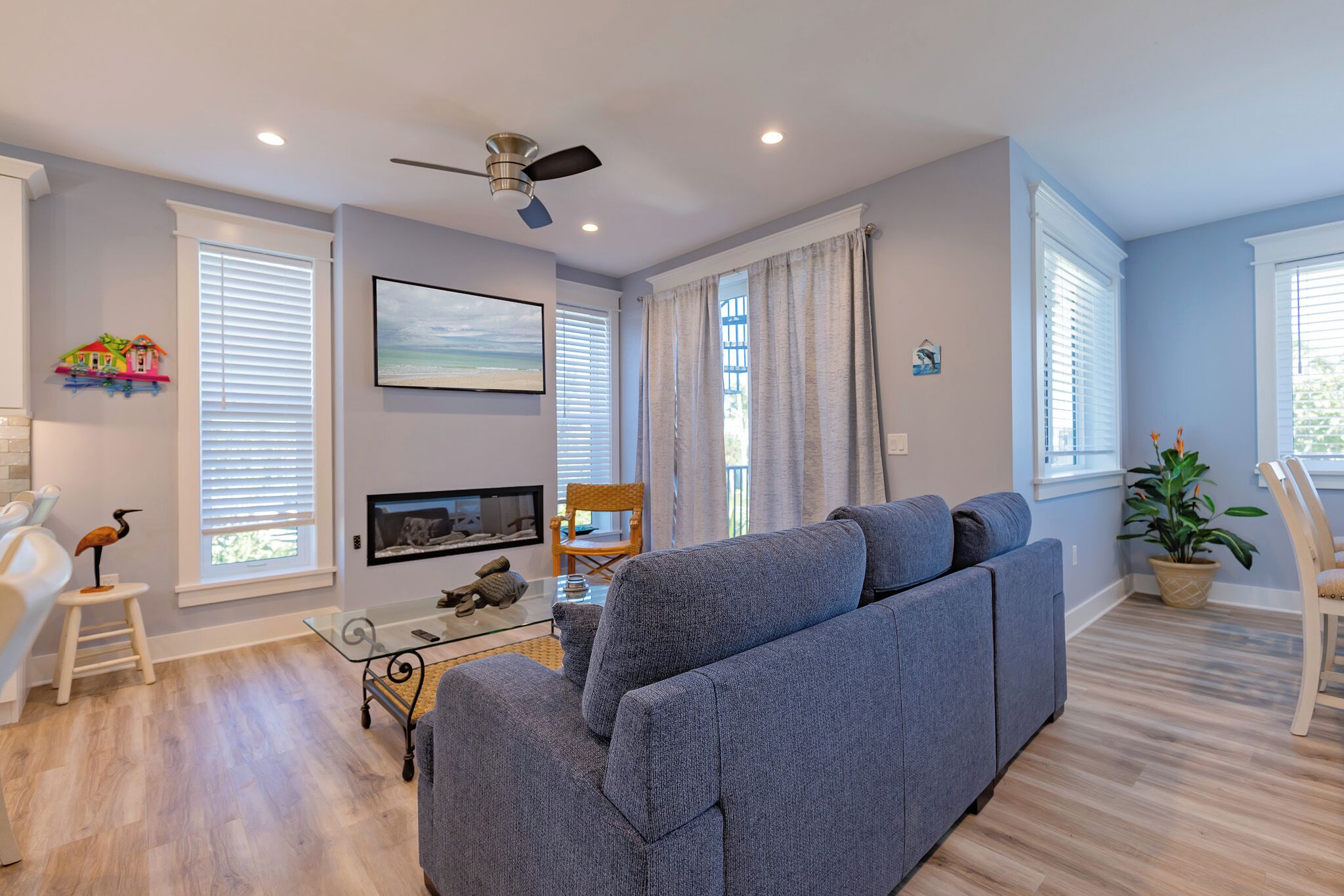 A modern living room with a grey sofa, a ceiling fan, a flat-screen TV, and a glass coffee table.