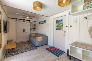 Ein kompakter Wohnraum mit einer Couch, einem kleinen Tisch, einem Deckenventilator und einer Tür mit einem farbenfrohen Poster.