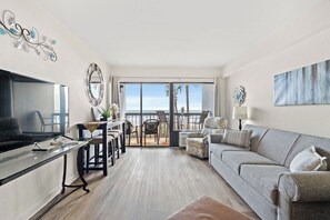 En rummelig opholdsstue med en stor sofa, et spisebord med stole, et fjernsyn og en balkon med havudsigt.