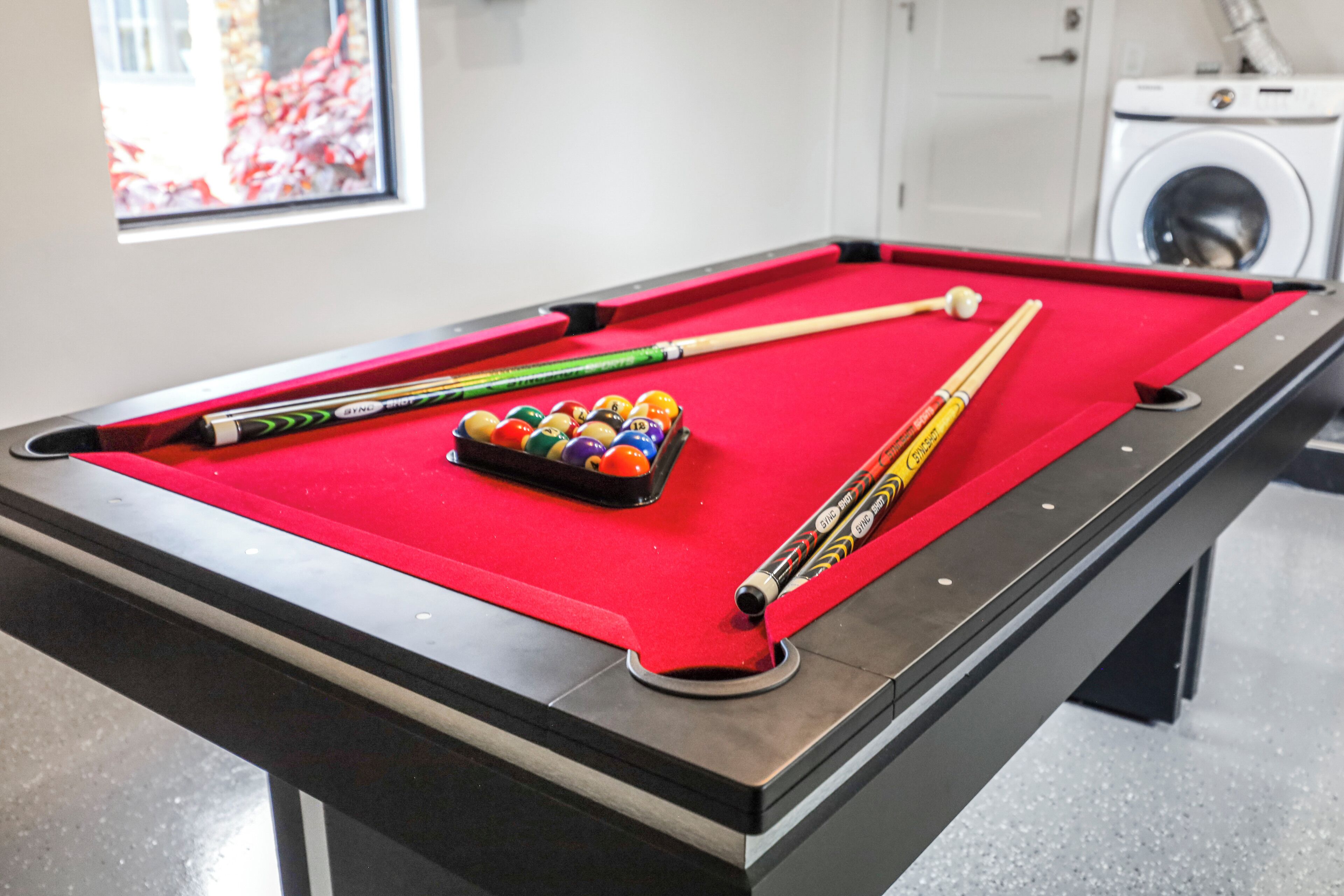 A pool table with balls and cues, a window with a view of red foliage, and a washing machine in the background.