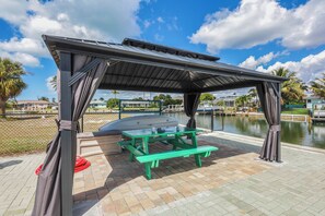 A covered picnic area with a table and benches, a pool in the background, and a fence surrounding the area. - Waterfront Home | Pool, Hot Tub, Kayak, & Billiards (Marco Island)