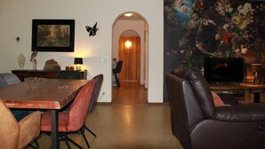 A dining area with a wooden table and chairs, a television, and a large floral painting on the wall. - Altstrimmig Shine (Altstrimmig)