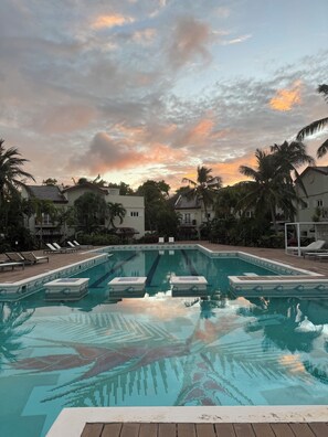 A swimming pool with clear water, surrounded by lounge chairs and palm trees, set against a backdrop of buildings and a sunset sky.