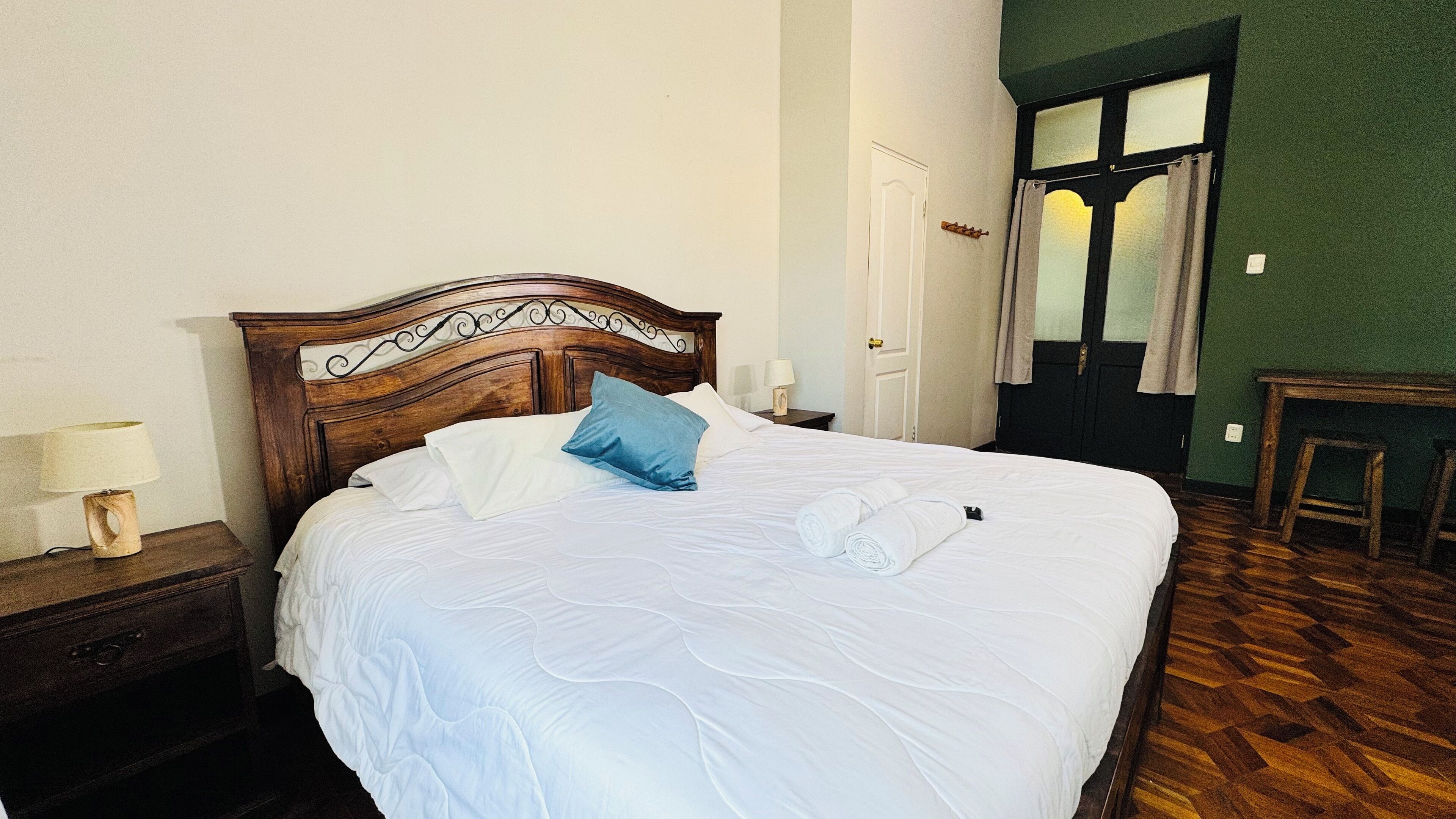 Cama bem arrumada com roupa de cama branca, cabeceira de madeira e travesseiro azul. Há uma mesa de cabeceira de madeira com uma luminária e uma pequena mesa ao lado da cama.
