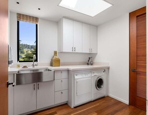 A compact kitchen with a built-in washing machine, a window with a view, and a wooden door. - Hedgerow Haven | Premium Oceanfront with Hot Tub (The Sea Ranch)