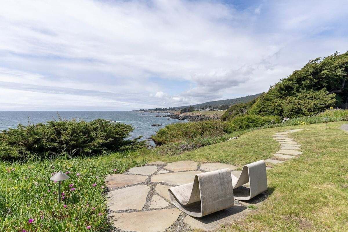 A coastal landscape with a stone pathway leading to a seating area with two chairs, overlooking the sea and a hilly backdrop.
