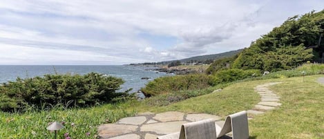 A coastal landscape with a stone pathway leading to a seating area with two chairs, overlooking the sea and a hilly backdrop.