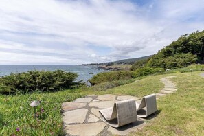 A coastal landscape with a stone pathway leading to a seating area with two chairs, overlooking the sea and a hilly backdrop. - Hedgerow Haven | Premium Oceanfront with Hot Tub (The Sea Ranch)