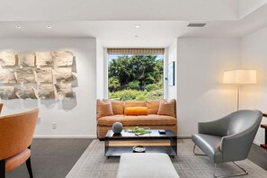 Ein modernes Wohnzimmer mit Sofa, Sessel und Couchtisch, einem Wandkunstwerk aus Stein sowie einem Fenster mit Blick auf Grünflächen.