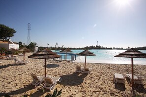 Uma praia com espreguiçadeiras e guarda-sóis, piscina e céu claro.