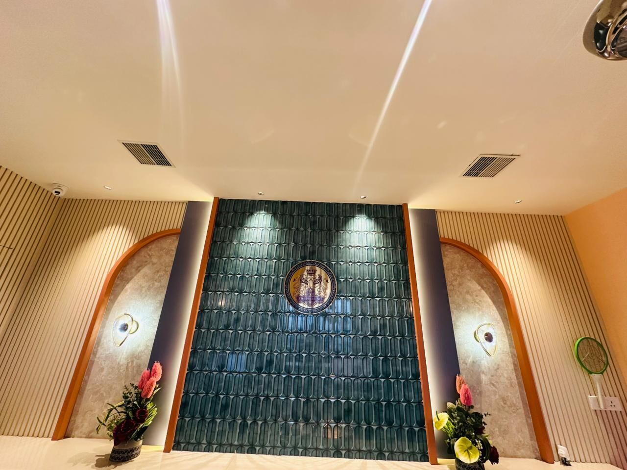 Un área de recepción con una pared decorativa, un mostrador de recepción y dos arreglos florales.