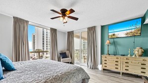 Una camera d'albergo con un letto, una cassettiera, una ventola a soffitto, un'ampia finestra e un quadro raffigurante una spiaggia.