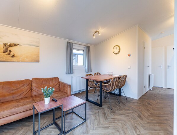 Uma sala de estar moderna com um sofá de couro marrom, uma mesa de centro de madeira, uma mesa de jantar com cadeiras de vime, um quadro na parede e uma janela com cortinas.