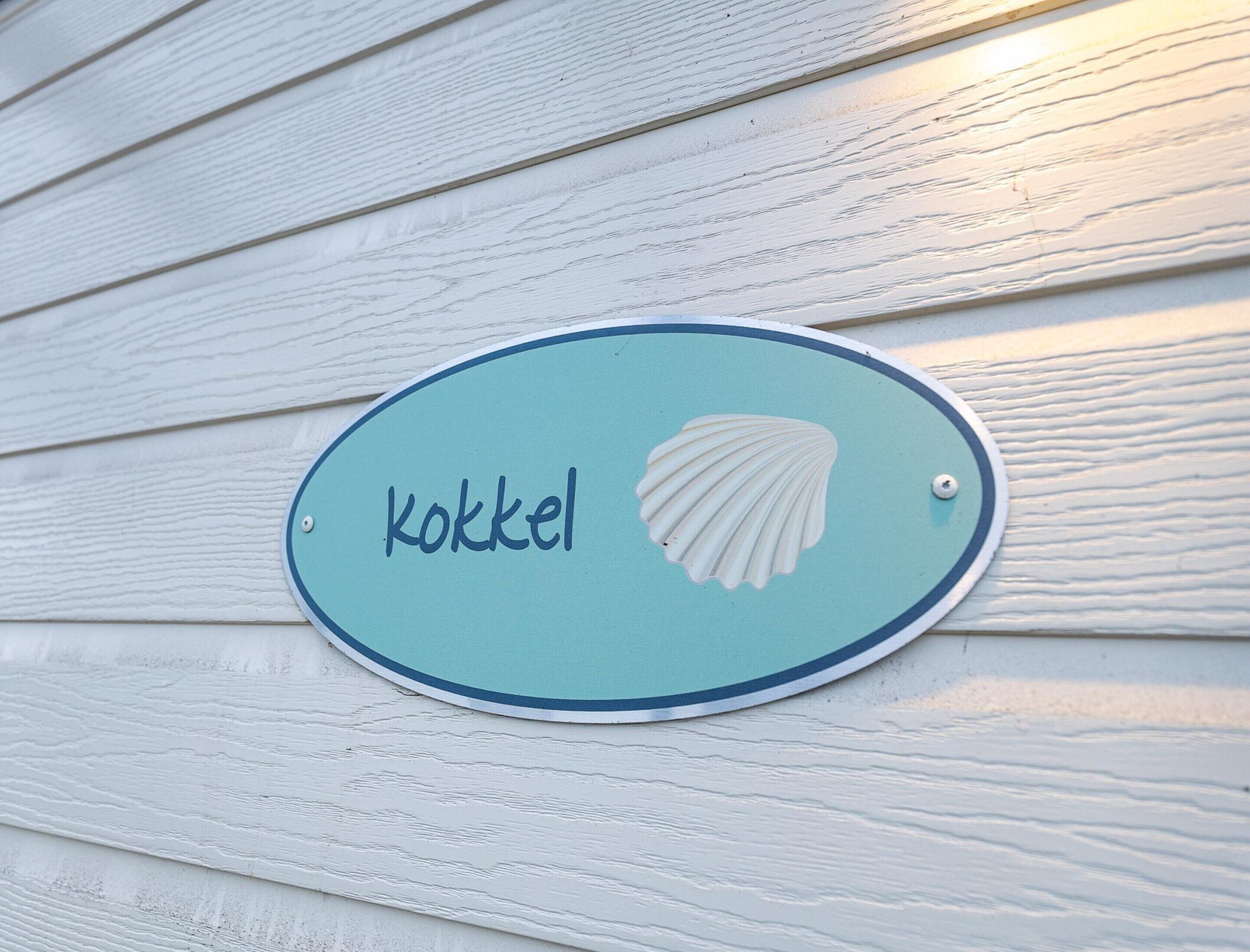Et skilt med ordet 'Kokkel' og en tegning af en snegl mod en turkis baggrund, monteret på en hvid trævæg.