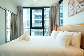 Ein Hotelzimmer mit einem großen Bett, einer Kopfteil und Blick auf eine Stadtlandschaft durch große Fenster.