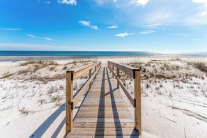 Ein hölzerner Steg führt zu einem Strand mit Sand und spärlicher Vegetation.