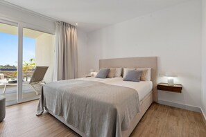 Ein modernes Schlafzimmer mit einem großen Bett, einem Nachttisch und einem Sessel neben einer Schiebetür zu einem Balkon.