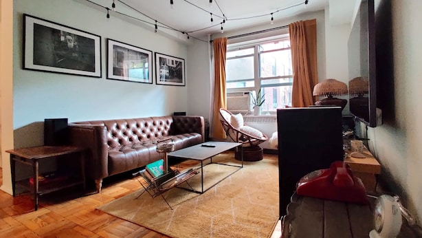 Un salón con un sofá de cuero marrón, una mesa de centro de vidrio, un sillón de mimbre y un televisor sobre un mueble bajo. Hay cuadros enmarcados en la pared y una ventana con cortinas.
