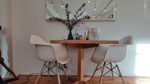 Un comedor con una mesa de madera, dos sillas blancas, un espejo sobre ella y luces colgantes.