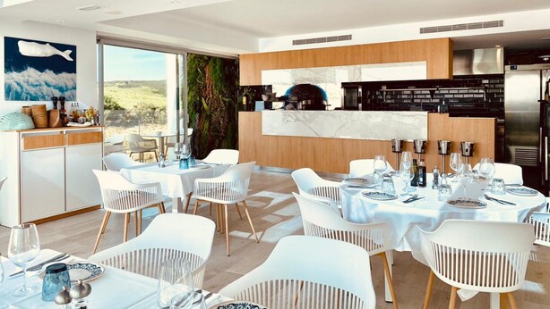 A modern dining area with white chairs and tables, a kitchen with a marble countertop, and a large window offering a view of the outdoors. - Exclusive 3-bed Penthouse Alcazaba Lagoon (Casares)