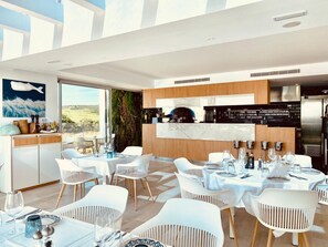 Zona pranzo moderna con sedie e tavoli bianchi, cucina con piano di lavoro in marmo e un'ampia finestra con vista sull'esterno.