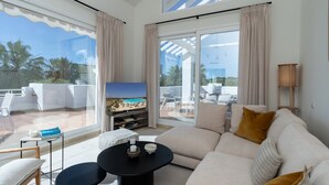A modern living room with a large sofa, a coffee table, and a TV. There is a balcony with a view of the beach and palm trees. - Exclusive 3-bed Penthouse Alcazaba Lagoon (Casares)