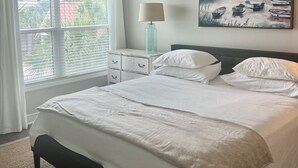 A bedroom with a bed, a ceiling fan, a window with curtains, a nightstand with a lamp, and a painting on the wall. - Delightful 2-bedroom renovated condo with AC in charming Charleston (Charleston)