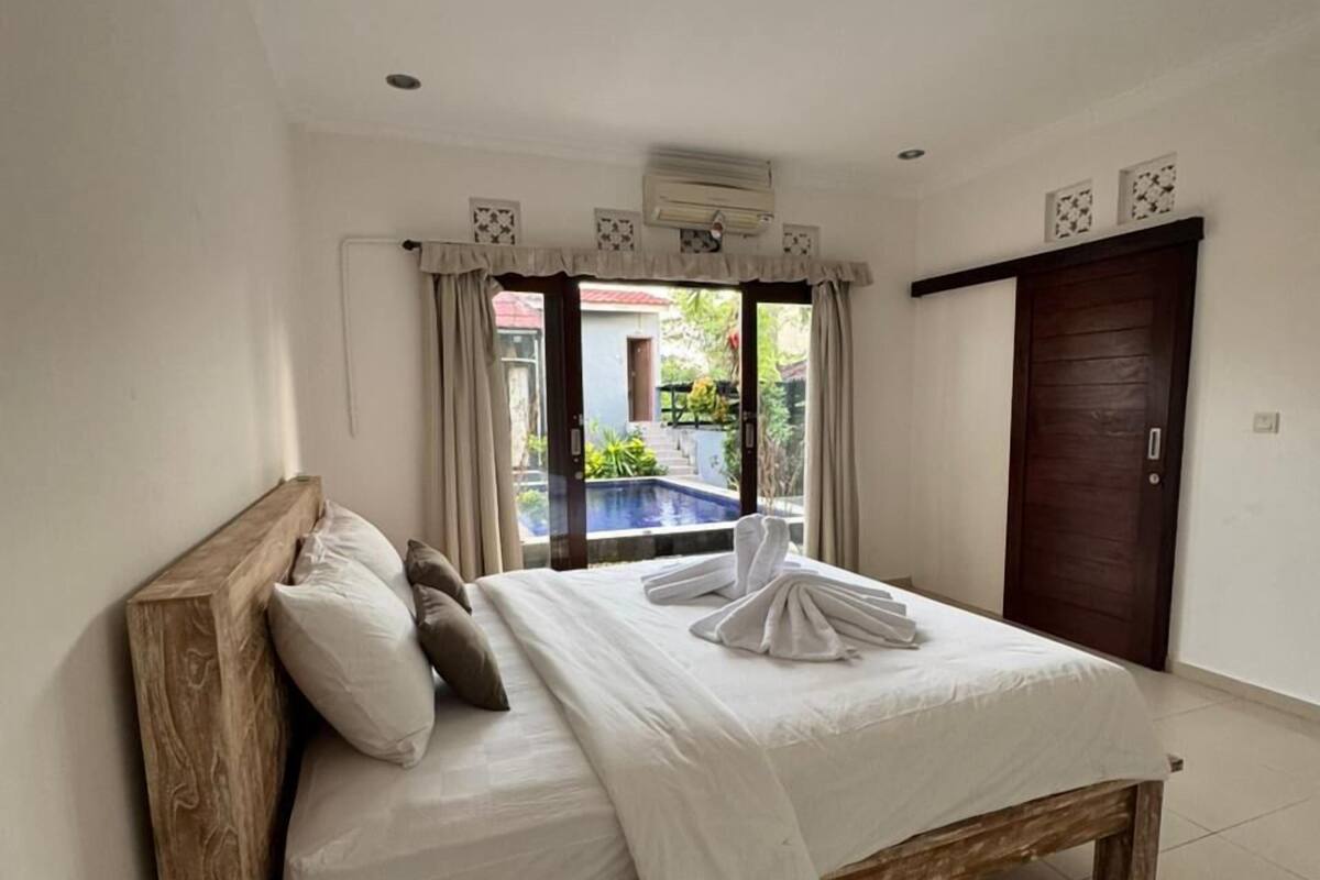 Een slaapkamer met een houten bed, witte beddengoed, uitzicht op een zwembad en een donker houten deur.