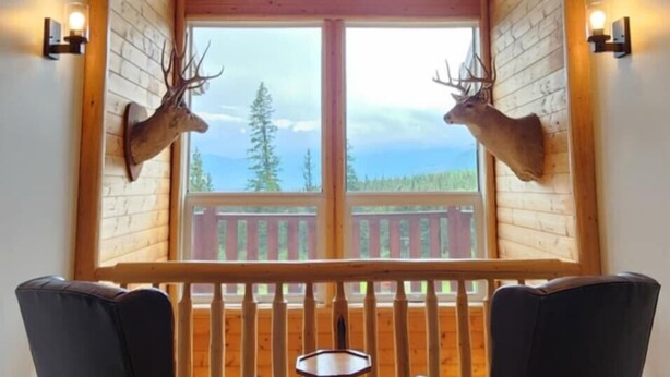 Una habitación con techo de madera, dos sillas, una mesita y un ventanal con vistas a árboles y montañas.
