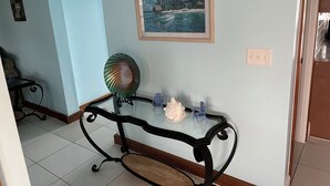 A room with a glass-top table, a decorative fan, a small statue, and a framed painting of a tropical scene.