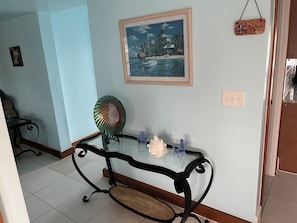 A room with a glass-top table, a decorative fan, a small statue, and a framed painting of a tropical scene.