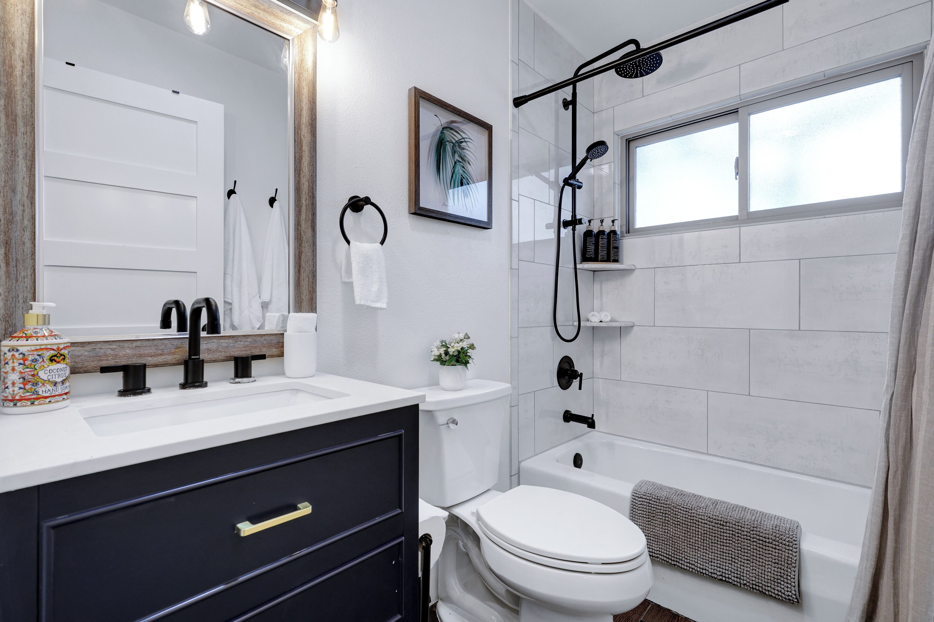 Ein modernes Badezimmer mit einem weißen Waschbecken, einem schwarzen Wasserhahn, einer Toilette, einer Dusche mit Glastür und einem gerahmten Bild an der Wand.