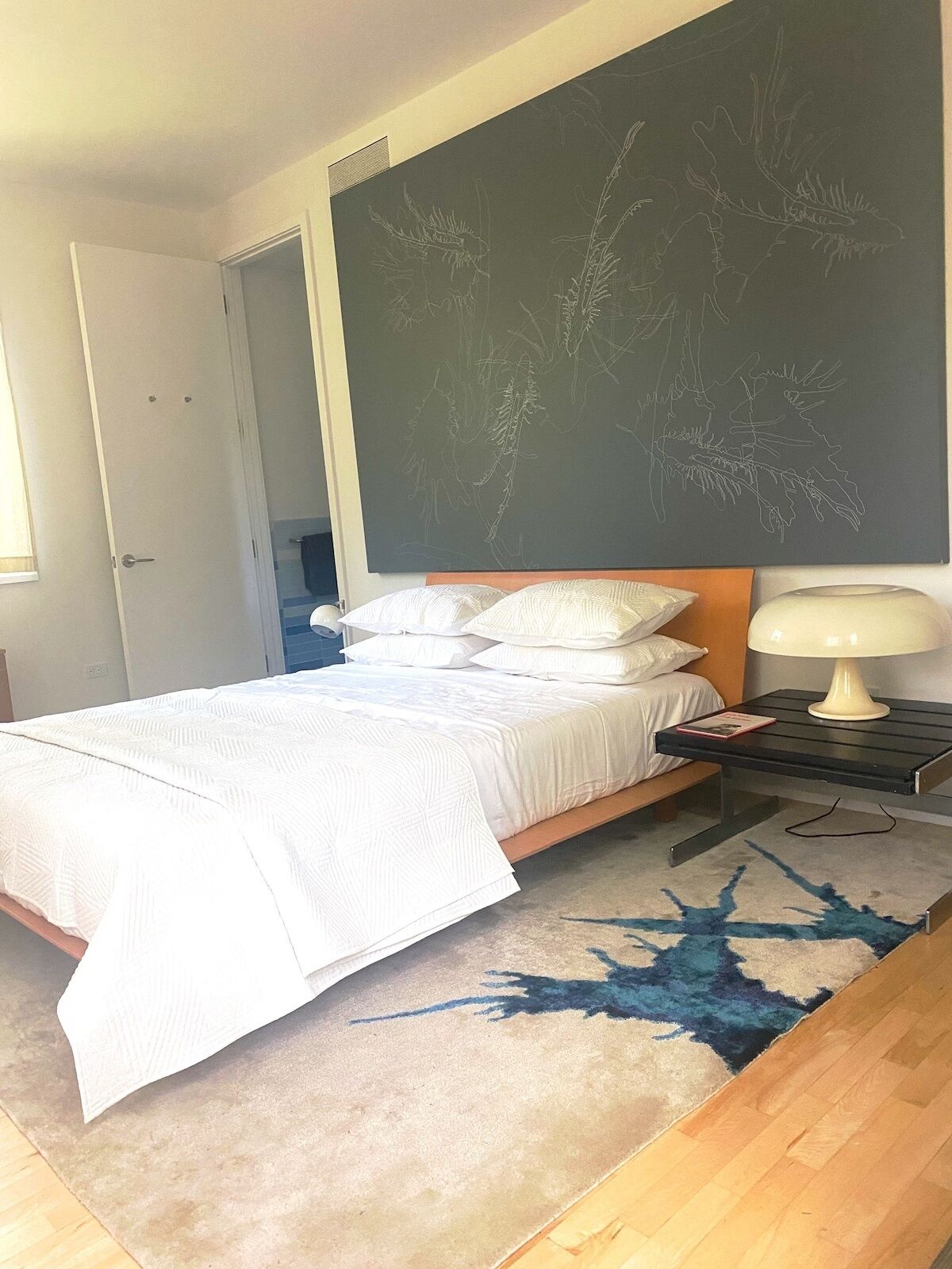 A bedroom with a bed, white bedding, a nightstand with a lamp, and a large chalkboard with plant drawings on the wall.