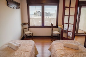 Ein Zimmer mit zwei Betten, einem Fenster mit Blick auf Gebäude, einem Stuhl und einem Kleiderständer.